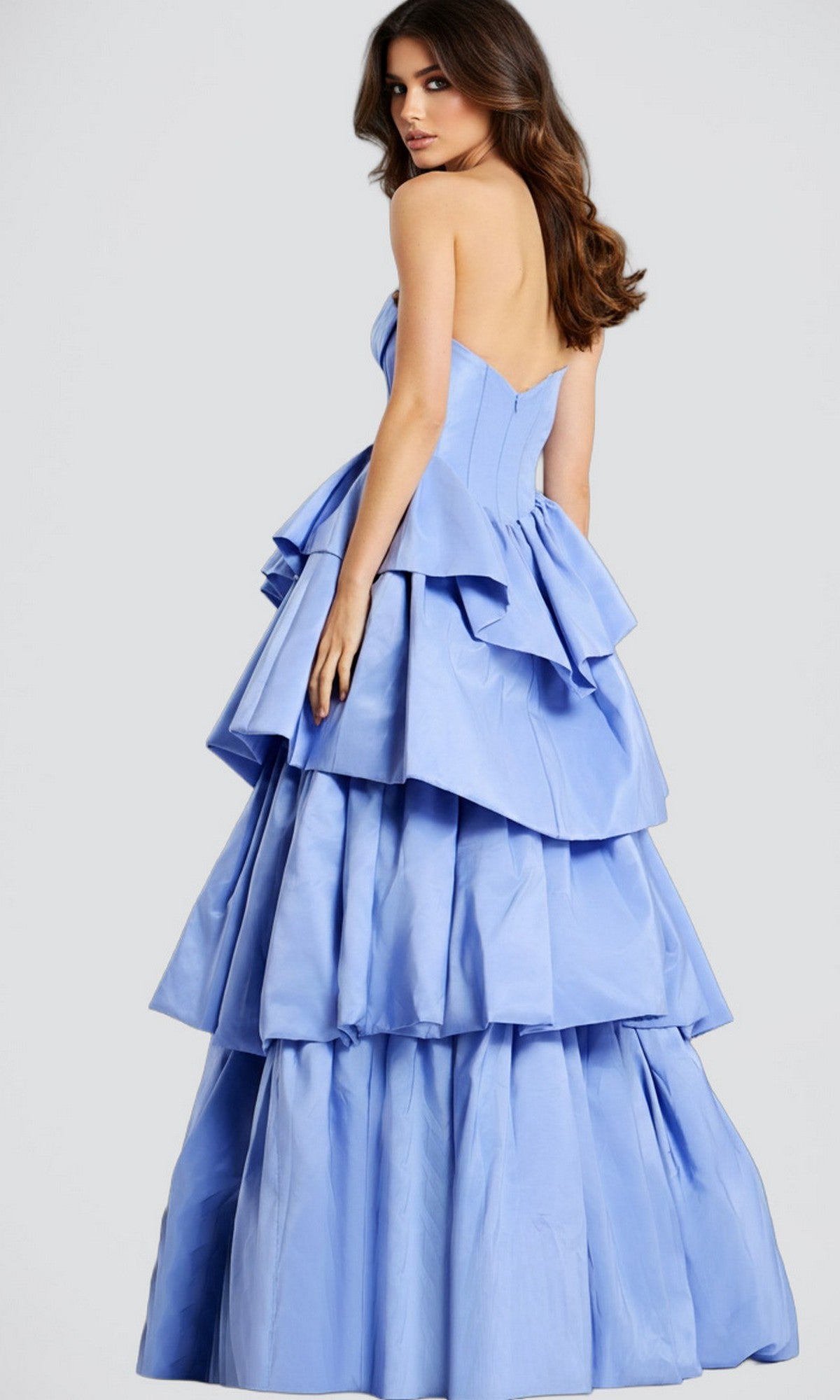 Woman wearing the Jovani 45111 Strapless Ruffle Long Prom Ball Gown in light blue, featuring tiered layers, poses looking over her shoulder against a plain background.